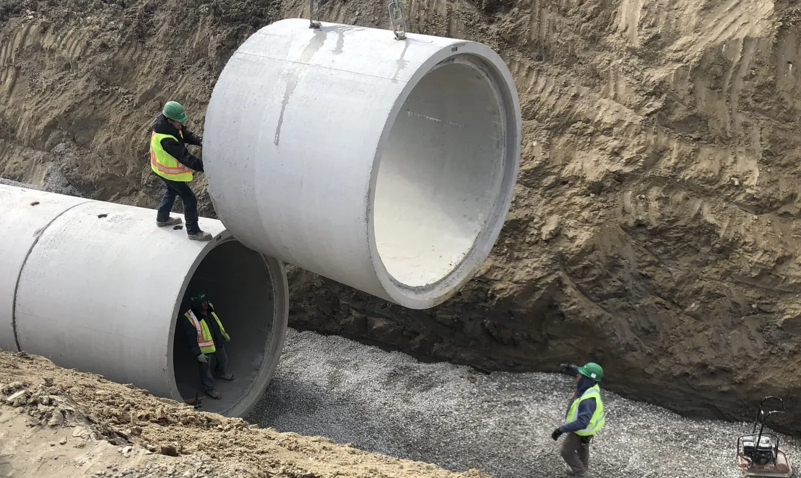 Workers guiding a large concrete pipe section into place during underground utility installation