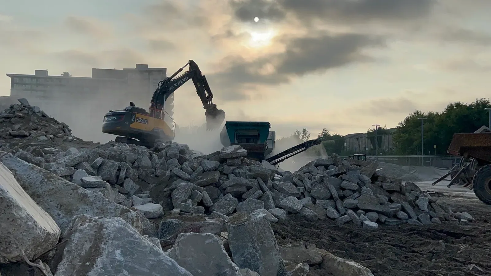 Excavator loading broken concrete into a crusher at an active construction site during early morning light.